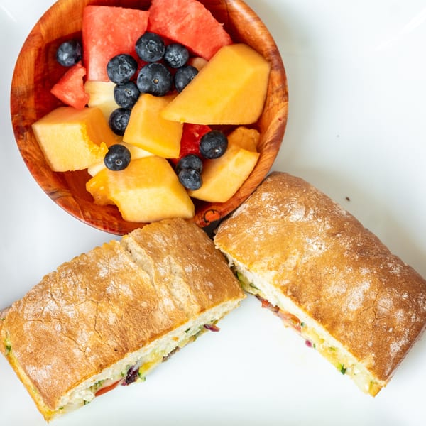 A ciabatta sandwich cut in half, served with a wooden bowl of fresh fruit salad.