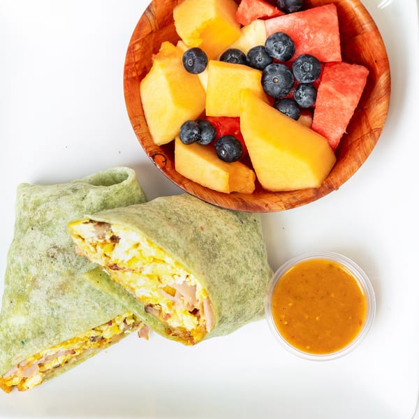 A green breakfast burrito, fruit bowl with melon and blueberries, and a side of sauce on a white plate.