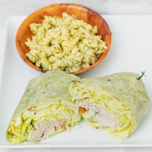 A green wrap filled with lettuce and tuna, cut in half, served with a side of pasta salad.