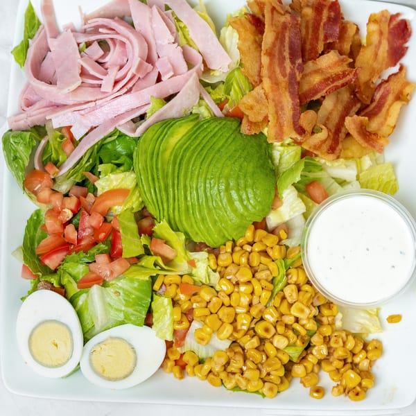A vibrant salad with ham, bacon, avocado, corn, tomatoes, hard-boiled eggs, and ranch dressing.