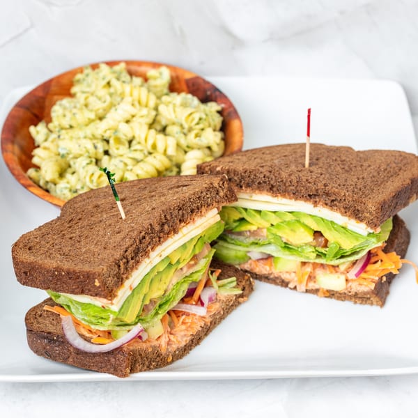 Two dark bread sandwiches with avocado, lettuce, and cheese, served with a side of pasta salad.