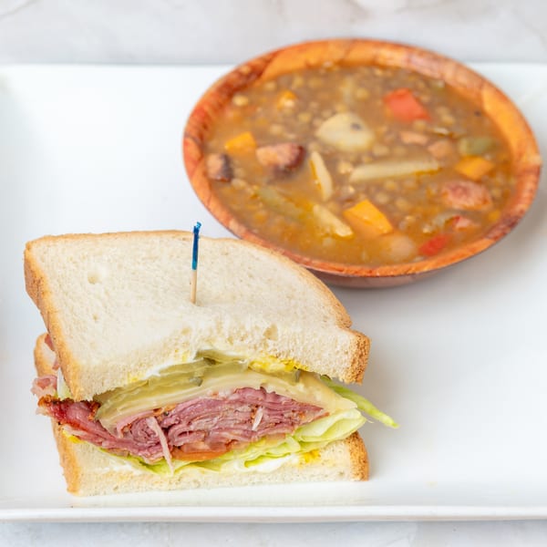 A deli sandwich with meat, cheese, lettuce, and pickles, next to a bowl of vegetable soup.